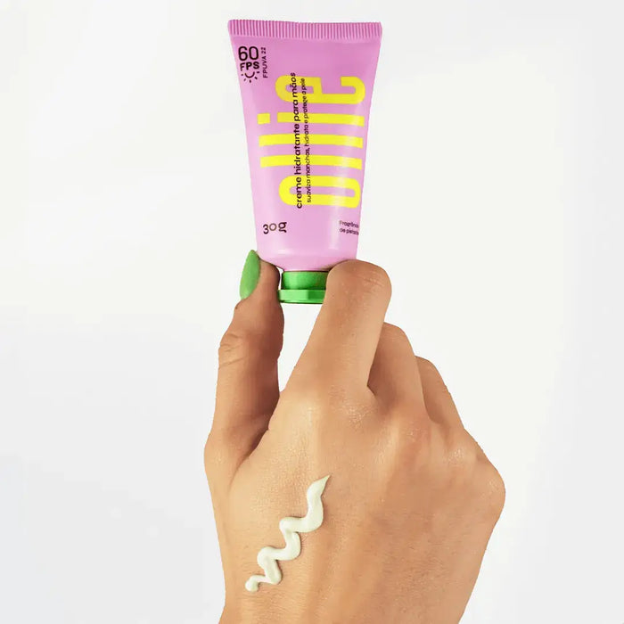 Mão segurando embalagem do creme hidratante para as mãos da Ollie pela tampa, enquanto apresenta um pouco do produto no dorso de sua mão.